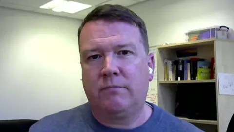 A man in a blue jumper wearing ear pods sits in an office in front of a bookshelf and looks into the camera