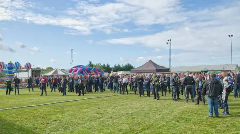 Will Badman Photography A group of people in a field with a set up for an event, including bouncy castle, gazebos and a fun fair ride