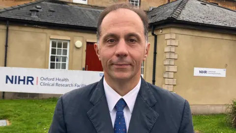 Professor Andrea Cipriani standing outside Oxford Health Clinical Research Facility. He has short dark hair and is wearing a grey suit with a white shirt and a blue tie. There is an NIHR Oxford Health Clinical Research Facility sign. The building is brown with grass in front.