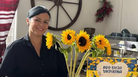 Yuliia Kryvenko A woman with dark-coloured hair is wearing a black shirt and is standing next to a bunch of sunflowers. There is a wooden wheel on the white-painted wall behind her. There is also a coffee machine in the background of the image.