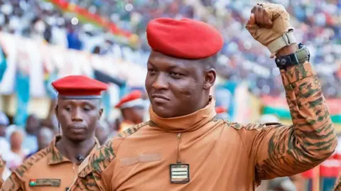 Ibrahim Traoré in beige military fatigues and red beret, surrounded by several soldiers in similar dress, pumps his fist in a stadium of his supporters.