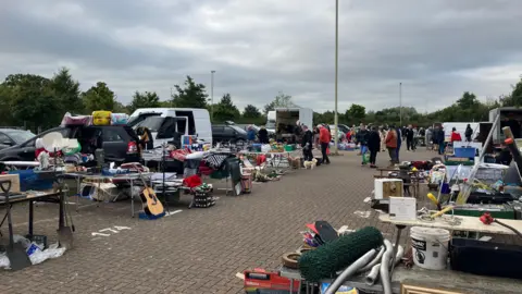 A car park full of car boot stalls selling various items from paint, blankets and spades to blankets a guitar and bric a brac. Half a dozen people are milling around having a look at the items in the middle distance of the photo.