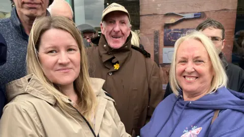 Two women satnd to the right and left of a man. The man is wearing a brown rain coat and light brown baseball cap. The lady to his right has light brown hair and is wearing a beige coat. The other lady is smiling broadly, has blond hair  and is wearing a blue jacket