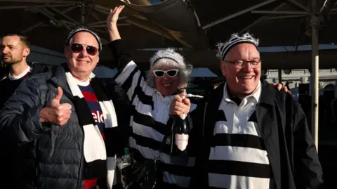 Michelle Mercer/Getty Images A woman with black and white sunglasses and a black and white top stands between two men with Newcastle United shirts.