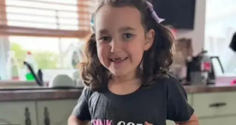 Family tells a smiling babe king showing off her t-shirt that has evil musical and title "Pink with green".