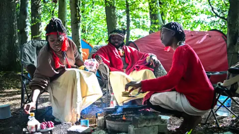 Getty Images Three people sitting on camp chairs in woodland. A large man in red and cream robes sits at the back. Two women are tending to a fire. There is a red tent in the background. 