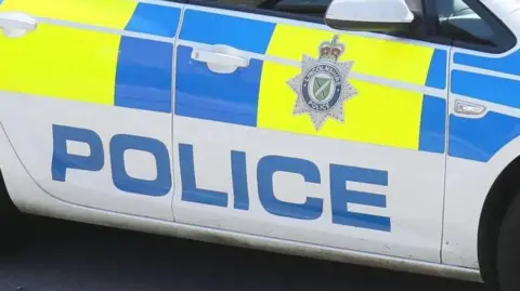 The side of a white police car, with blue and bright-yellow decor. The word "police" is printed in large blue letters and a Lincolnshire Police badge is fixed to the door.