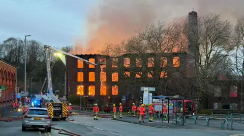 Carl Haslam/GMFRS Daylight view as crews contine to tackle the orange flames inside the three-storey brick mill, which has a tower. Firefighters can also be seen on the road.