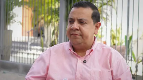 Javier Tarazona sits in a garden wearing a pink shirt looking serious