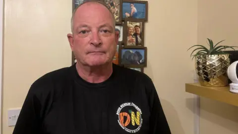 Mark Nickolay A man with a glum face, wearing a black shirt with the Donna Nickolay Foundation logo on. He is stood behind a series of pictures of his late daughter Donna inside his house