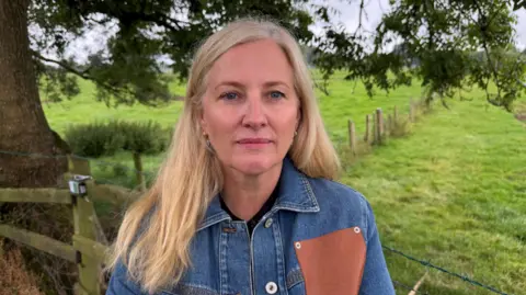 Sarah Herbison has blonde hair and a denim jacket with a brown quare on it. She is standing in front of a field with part of a tree behind her.
