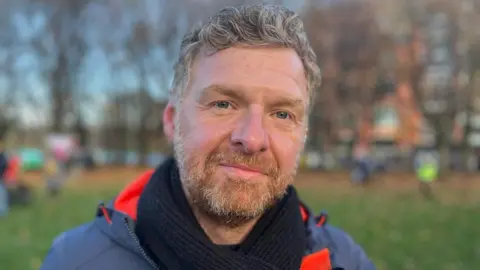 A man with short curly light brown hair and a beard is standing outside in a park. He is wearing a dark scarf and an orange-lined jacket. Trees and people are blurred in the background.