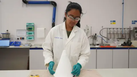 A woman with black hair is wearing safety goggles along with a white lab coat and green plastic gloves. She is working in what appears to be a laboratory.