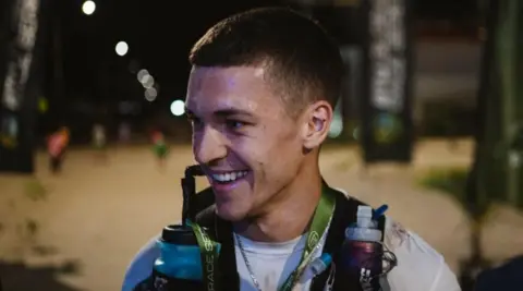 A man is smiling looking side on to the camera in the dark. He is wearing a white top.
