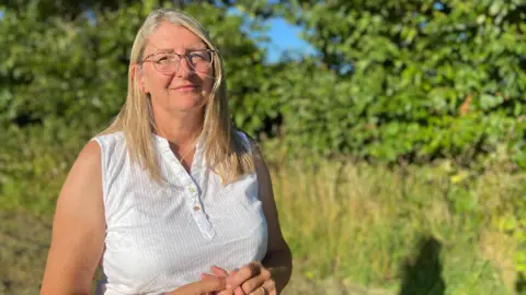 A woman with long blonde hair is standing outdoors in a sunny area with green trees and bushes in the background. She is wearing a sleeveless white shirt with buttons at the top. She has her hands clasped together in front of her.