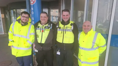 North Tees and Hartlepool NHS Foundation Trust Maintenance assistant Luke Ferguson, security team members Gary Pickard and Aaron Sparrow, and plumber Ste Loughran smiling outside the University Hospital of North Tees. They are wearing high visibility clothing.