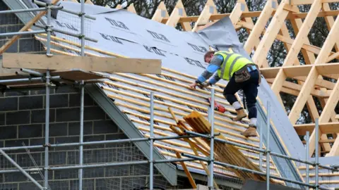 PA Media A construction worker works on a roof of a new house that is being built. Scaffolding can be seen in the foreground and the wooden frame of a roof of a house in the background.