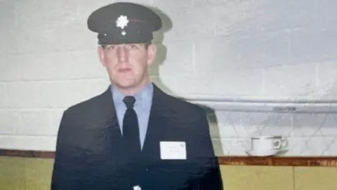 Handout A photograph of Jim McNeil when he was younger. He is looking into the camera and wearing a dark uniform and cap. A cup and saucer is on a wall behind him.