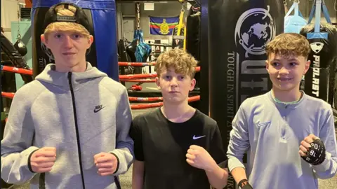 Three young boys standing in the gym holding their fists up in a boxing stance