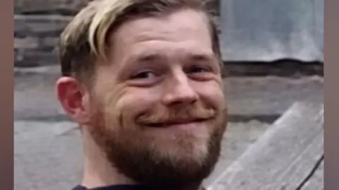 A man in his late 30s, with blonde hair combed to the side and a ligh brown beard, smiles at the camera. He is wearing a blue hooded jumper. 