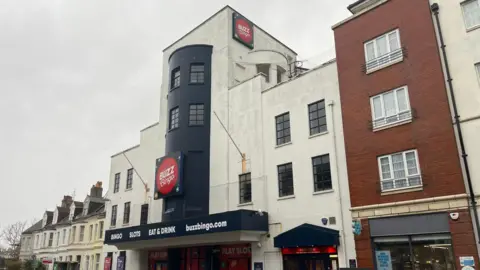 George Carden/BBC The Buzz Bingo building is an art deco style cream building which is four storeys tall. It has a large buzz bingo red sign on the front. There are cars parked outside the front