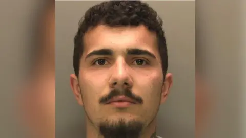 A man with short dark hair, slightly curly, with a black moustache and goatee beard is looking straight into the camera of a police custody picture.