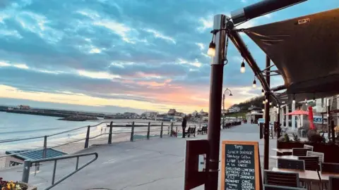 Tom's Lyme Regis Empty tables under a canopy on the promenade outside Tom's Lyme Regis restaurant. In the distance is the beach and harbour wall and a colourful sunset sky.