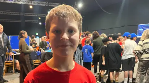 A boy with light brown hair, wearing a red t shirt, looking into the camera and smiliing. Behind him there are some children and teachers gathered - around 20 of them- most of them wearing trousers, t shirts and caps. They are huddled together talking. They are in a big space with black walls and silver lighting rigs on the ceiling - the Royal Jersey Showground.