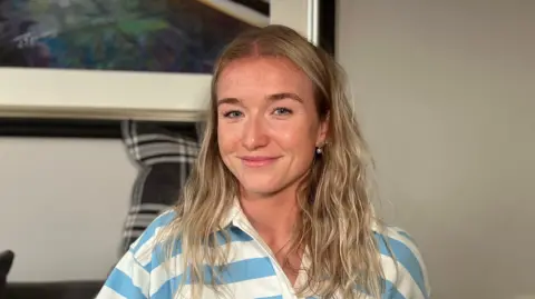 Lucy Woodward sits on a grey sofa indoors and is wearing a blue-and-white striped pullover, with framed artwork visible on the wall in the background.