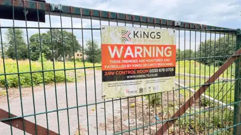 Phil Towers/BBC A section of land which is guarded by a tall metal gate. On the gate is a sign for the security company, which reads 'Warning - these premises are monitored and patrolled by Kings Security personnel'.