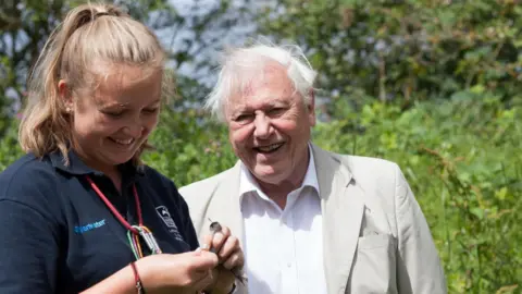 Eric Renno Sir David Attenborough pictured smiling with a member of staff from Leicestershire and Rutland Wildlife Trust. He is wearing a beige jacket and a white shirt. The member of staff is wearing a navy polo top and is also smiling. They are pictured on a sunny day with green foliage behind them. They are pictured at the opening of a volunteer training centre at Rutland Water Nature Reserve in 2015.