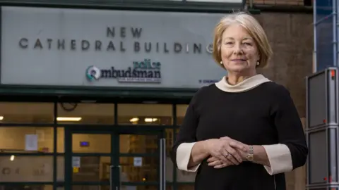 PA Media A woman in a black dress standing with her hands clasped together in front of a building that has "New Cathedral Buildings, Police Ombudsman" written above the door.