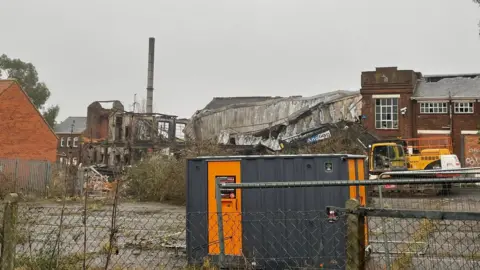 Chris Goreham/BBC Demolition plant at the site of the former factory. Part of the roof has collapsed and the ruins can be seen in the background 