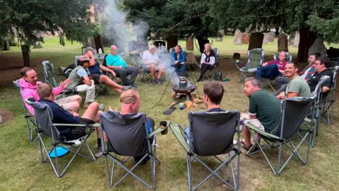 Jonathan Toney A group of men sit in camp chairs around a fire pit with smoke coming out of it - and a kettle sits on top of the fire.