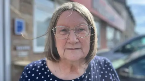 A close-up of Judith Robbins, who has straight, shoulder-length hair. She is wearing glasses with thin frames and a blue and white polka dot top. The blurred background features a building and two parked cars.