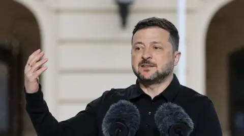 Volodymyr Zelensky speaking at a lectern with his hand raised
