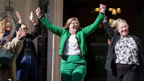 Following the ruling in the Supreme Court that the term woman refers to 'biological women', campaigners Susan Smith (in green suit) and Marion Calder, both directors of 'For Women Scotland' celebrate the judgement outside the court, on 16th April 2025, in London, England. Susan is holding both arms in the air and clasping the right arm of Marion who is in a black suit. Both women have light brown/blonde hair. Two other women to the left of the image are applauding.