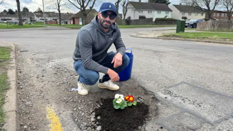 A man sitting in the road with a pothole in front of him which is filled with soil and flowers. 