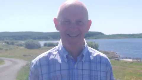 Maynard Cousley smiling at the camera, he is wearing a blue and white check shirt. Behind him it is sunny and there is a reservoir