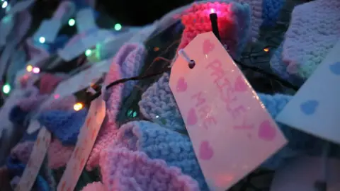 Knitted ribbons and name tags. One tag can be seen with the words: "Paisley-Mae" on it, surrounded by pink hearts  