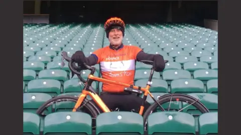 Patrick McIntosh A man in an orange t-shirt and long black sleeve shirt is sitting inside Twickenham stadium.