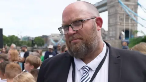 Joshua Falcon/BBC A head and shoulders picture of a man wearing glasses and a suit with a black blazer over a white shirt with a striped tie. Tower Bridge is in the background.