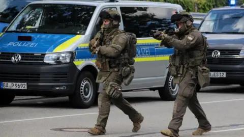 Special police walks down a road towards a suspected hostage-taking situation in a bank in Regensburg. A police van and other vehicles are parked in the background.