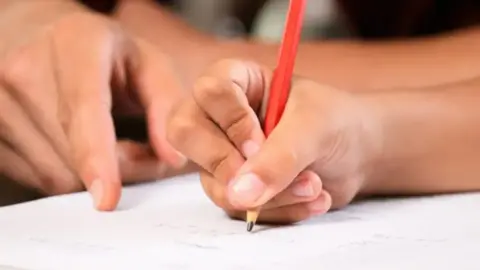 A child's hand holding a red pencil while an adult points at a white paper.