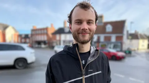 Stuart Woodward/BBC Jack smiles at the camera while standing in the centre of a market town or village, with roads and traditional buildings behind him. He wears a navy T-shirt with a thin running top zipped up over it.