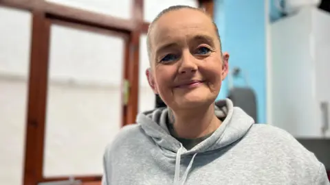 Emma Baugh/BBC Jo Evans is looking directly at the camera and smiling. She has her hair pulled back into a ponytail and is wearing a grey hooded sweatshirt.