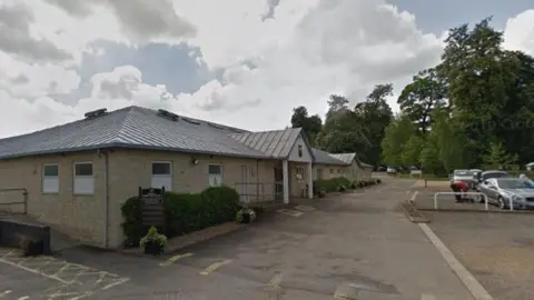 Google A single storey late 20th Century yellow brick building with a grey pitched roof is on the left. On the right is a car park with cars and beyond it mature trees in summer leaf. 