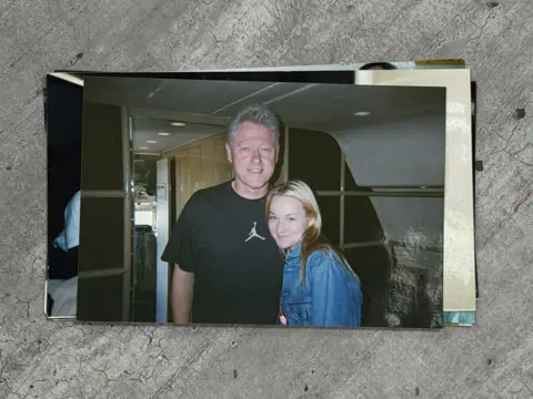Chauntae Davies Bill Clinton, an older man with short grey hair, stands close to a young woman, Chauntae Davies. Clinton is in a black short-sleeve t-shirt that has the Air Jordan logo on it. Davies is wearing a light-washed jean jacket. They are inside a plane.