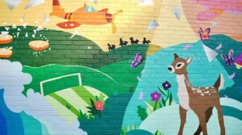 Part of a mural on a brick wall. It has rolling hills that are green, with splashing blue waves below it. There are purple butterflies flying and a brown deer in the foreground. The sky is pink and yellow and there is an orange plane flying across it.