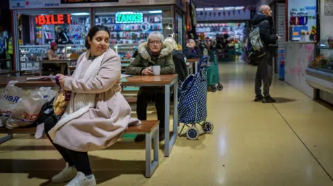 Getty Images People do their shopping in Gorton Market on January 28, 2026 in Gorton, United Kingdom. A woman can be seen sat on a bench with her shopping while an older woman sits with a cup of tea behind her. Others can be seen looking at stalls. 